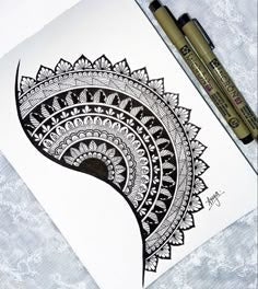 An intricate crescent-shaped mandala features detailed concentric bands filled with geometric patterns, floral motifs, and fine linework. Two black fineliner pens rest diagonally on the top-right corner of the paper.